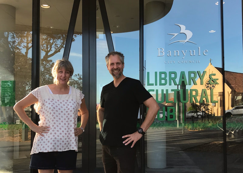 Smiling woman in white blouse, man in black tshirt, standing outside in front of glass wall that says Library.